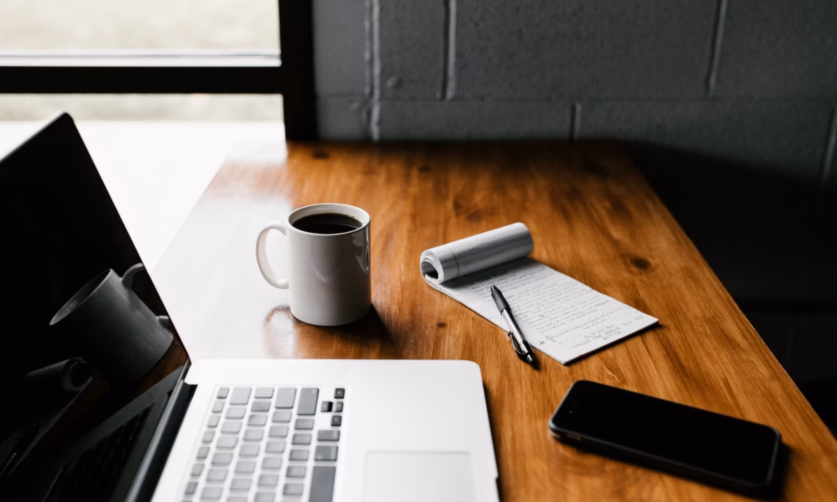 A real estate agent's laptop sits on a wooden table next to a cup of coffee, smartphone and pad of paper as the agent wonders what is email marketing and how can i use it to boost my business?