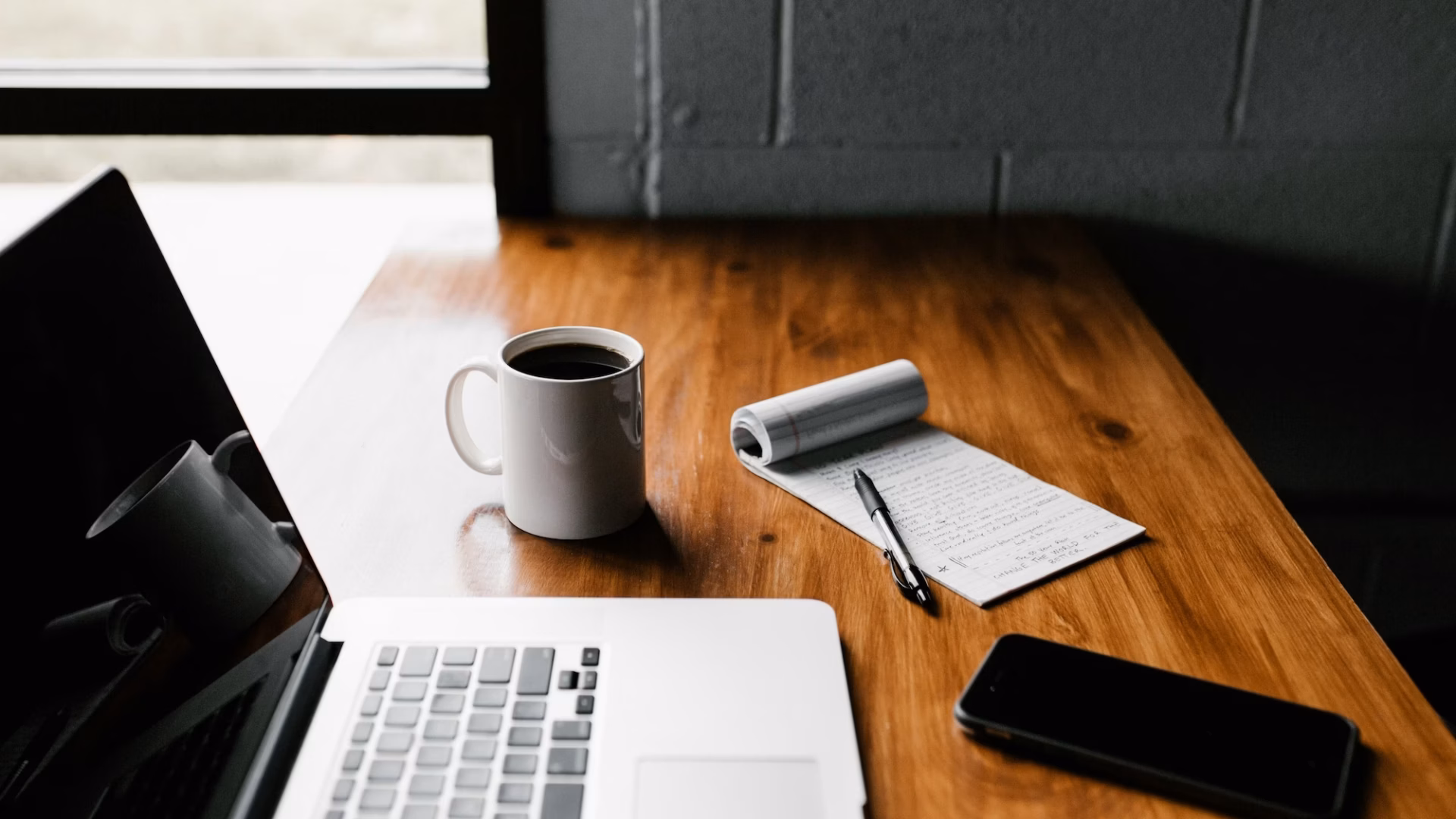 A real estate agent's laptop sits on a wooden table next to a cup of coffee, smartphone and pad of paper as the agent wonders what is email marketing and how can i use it to boost my business?