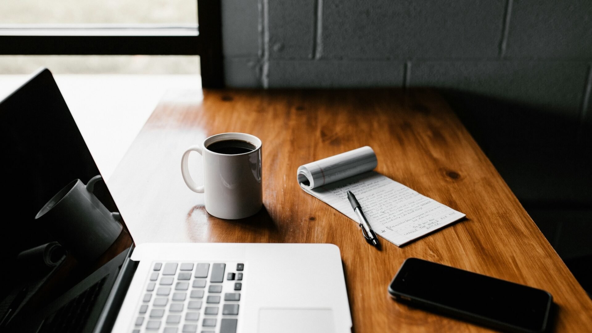 A real estate agent's laptop sits on a wooden table next to a cup of coffee, smartphone and pad of paper as the agent wonders what is email marketing and how can i use it to boost my business?