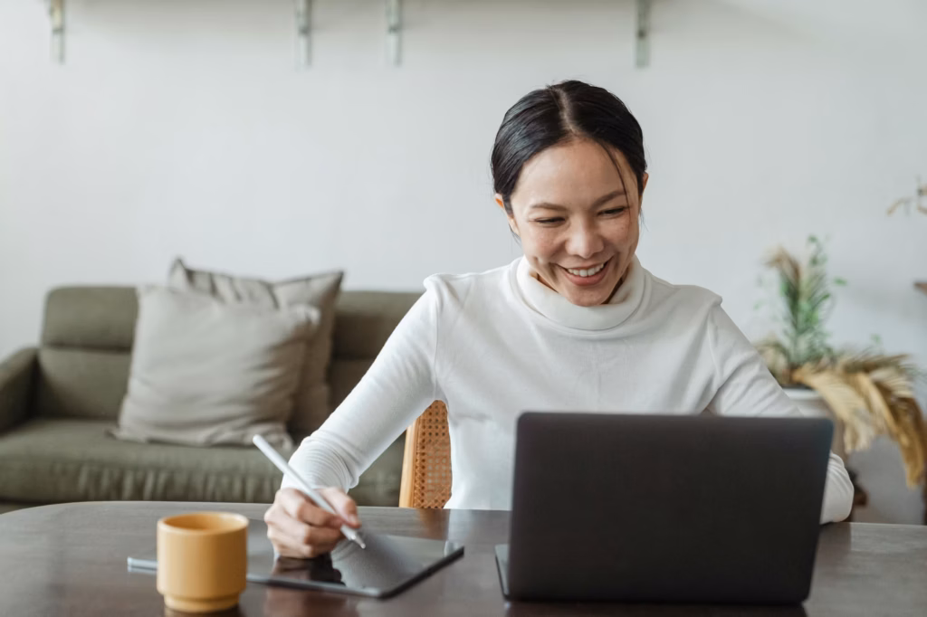 A real estate agent sits in front of a laptop smiling, jotting something down on her tablet about her Real Estate Marketing ROI with luxury presence