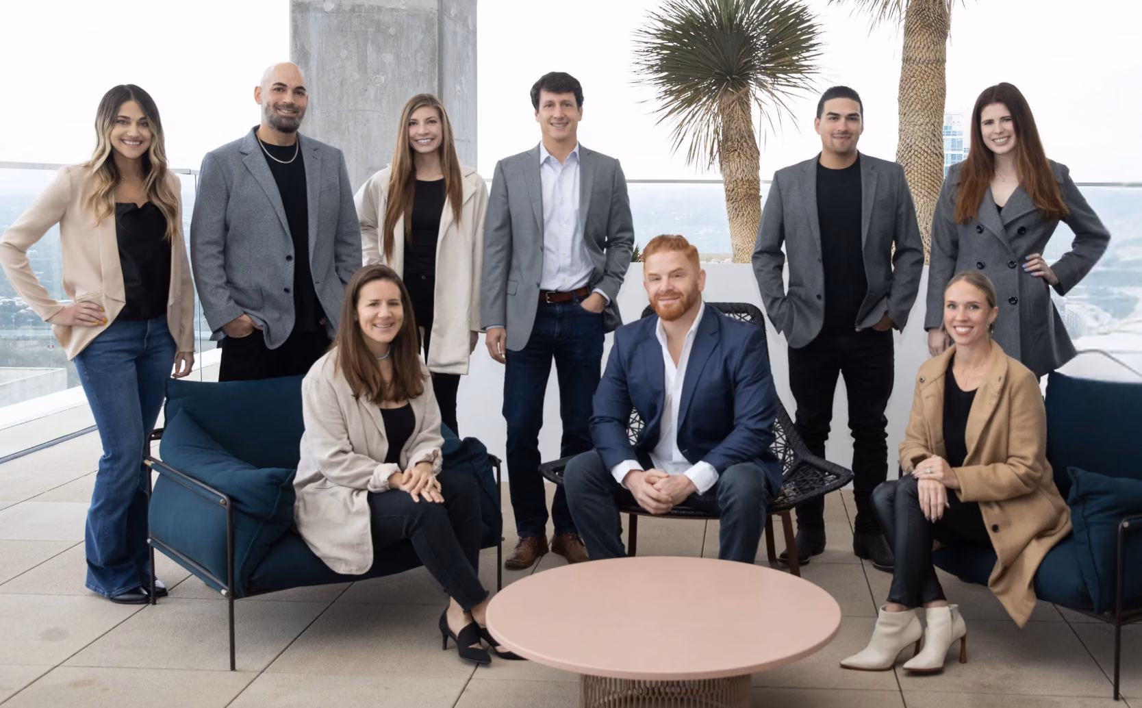 Real estate agents, brokers and support staff from The Zell Team in Austin, TX stand together in front of windows and palm trees for a group photo
