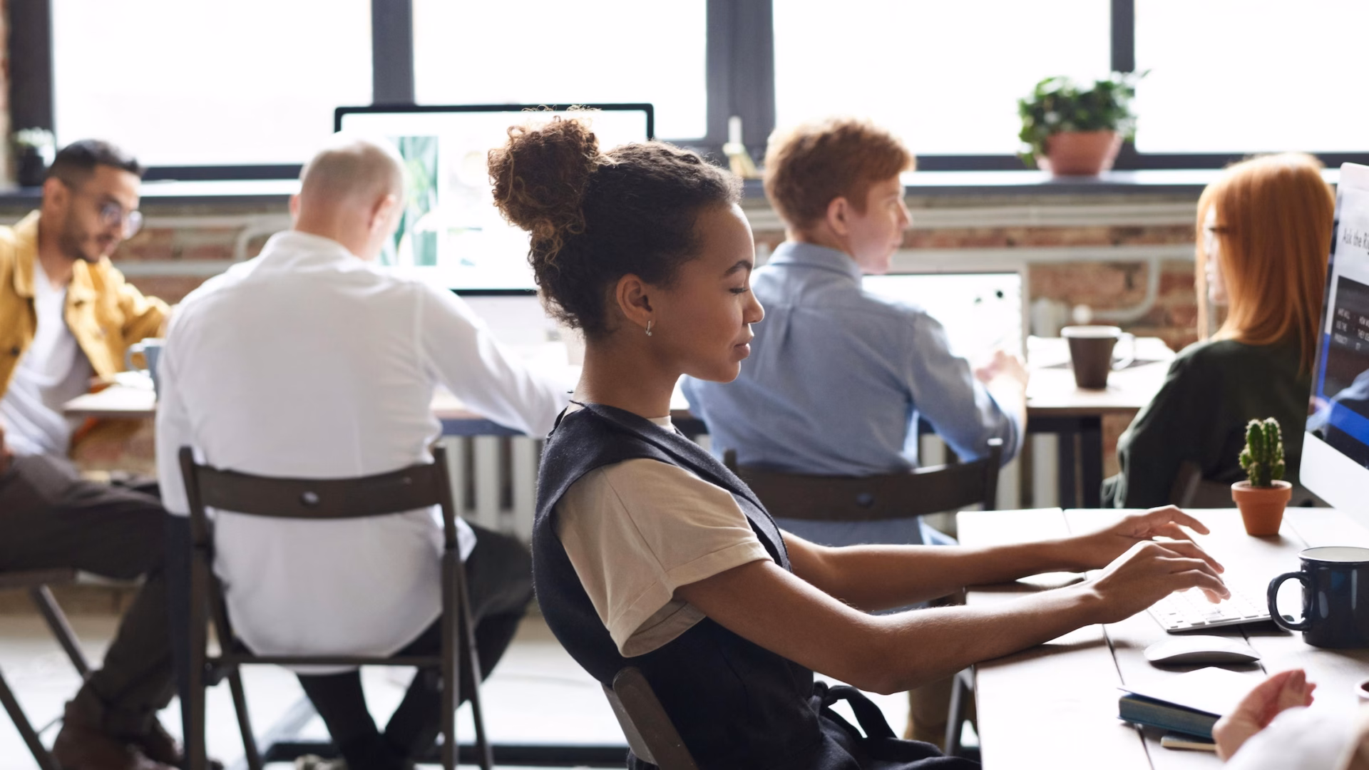 A real estate agent sits in front of a desktop computer in an office of her peers trying to decide which is the right platform between Luxury Presence vs. Real Geeks