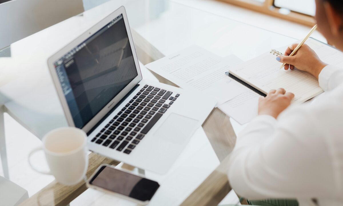 Real estate agent sitting at a desk in front of a laptop and cup of coffee, writing down pros and cons of Luxury Presence vs. Agentfire
