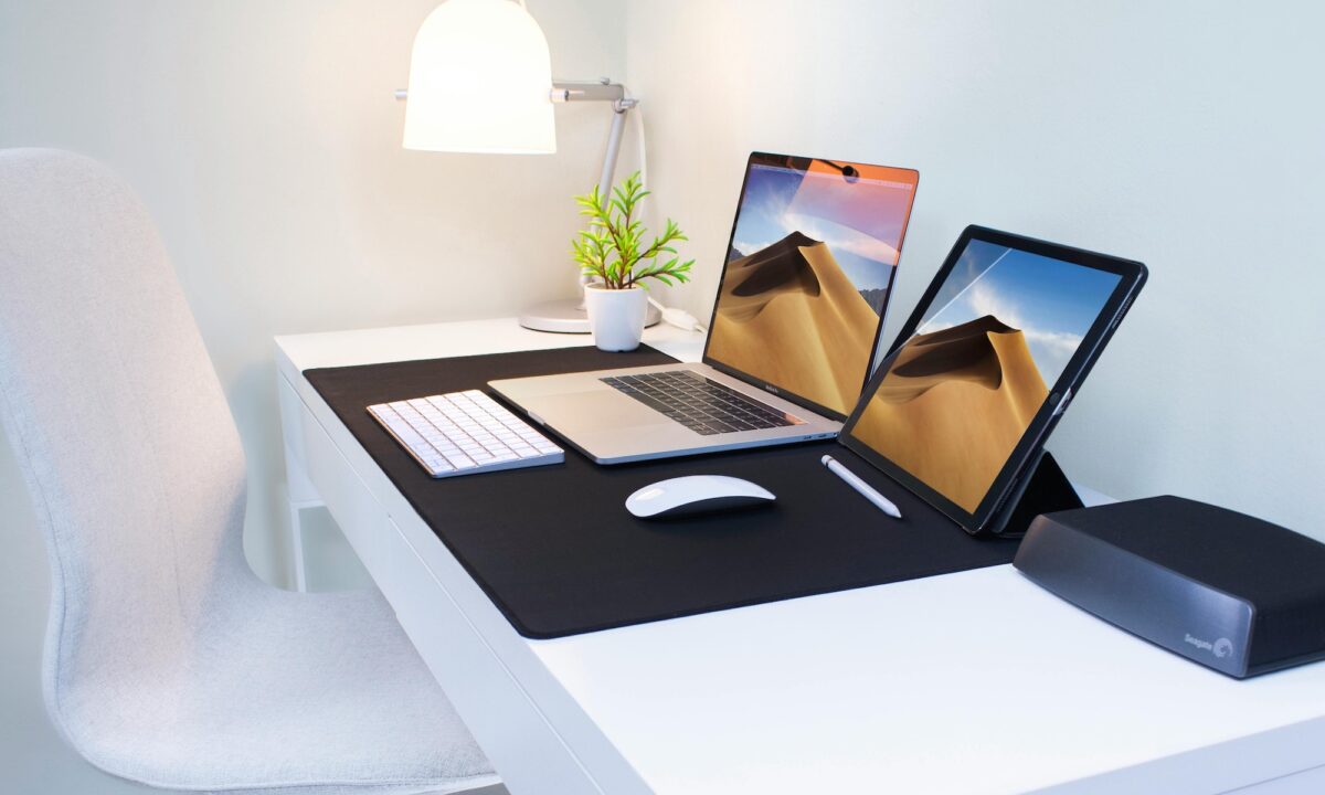 A white chair in front of a desk that has a laptop, tablet and lamp on it